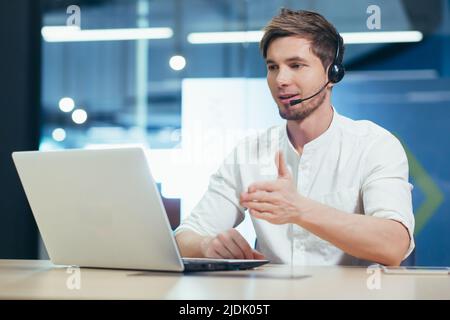 Video conference. A young handsome man in headphones sits in the office behind a laptop and communicates through a video call. Stock Photo