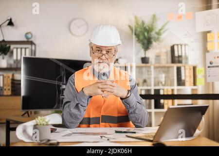 Close up portrait of mature builder drawing renovation house equipment architects on the desk with crossed fingers using blueprint in the office. Concept of architects, construction, design. Stock Photo