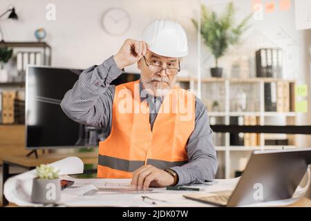 Professional senior male architect builder in white protective helmet and uniform using new digital laptop computer, working on construction plan, using sketches at his workplace. Stock Photo
