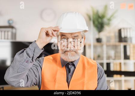 Portrait of mature architect wearing white construction helmet indoors. Bearded man in orange reflective vest posing among architectural office of the company. Stock Photo