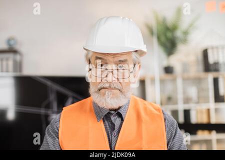 Portrait of mature architect wearing white construction helmet indoors. Bearded man in orange reflective vest posing among architectural office of the company. Stock Photo