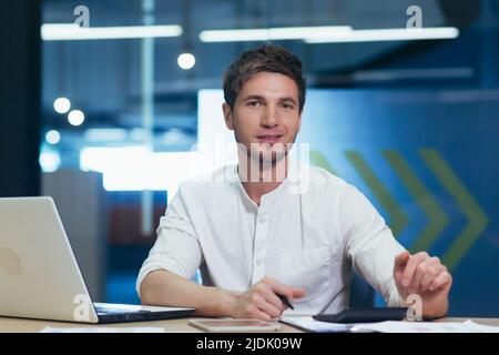 Portrait of a young handsome man sitting at a desk in the office in a white shirt. He looks at the camera, smiles Stock Photo