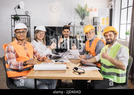 Qualified architects, engineers and designers having business meeting at office in front of TV screen. Multicultural colleagues in suits and helmets sitting at table and looking at camera. Stock Photo