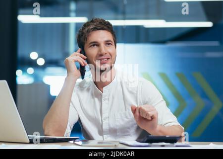Young handsome man, specialist working in the office at the desk. Talking on a mobile phone with customers, with bank, with business partners Stock Photo