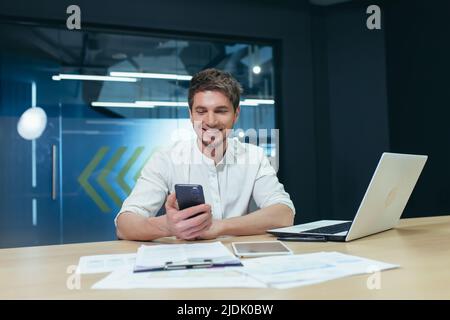 Young man, businessman, manager sitting in the office at his desk, holding a mobile phone, typing messages, making phone calls. Works on laptops and d Stock Photo