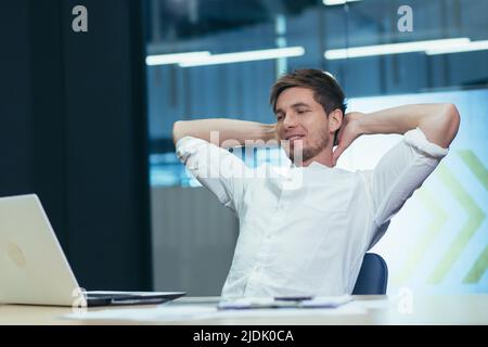 Young handsome man freelancer, businessman, manager in the office at the table resting, doing work during a break, throwing his hands behind his head, Stock Photo