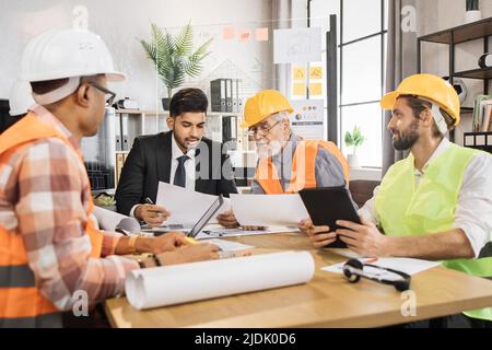 Multiracial men builders in formal outfit and helmets sitting at table with lots of blueprints during important strategic session. Business people working on common construction project. Stock Photo