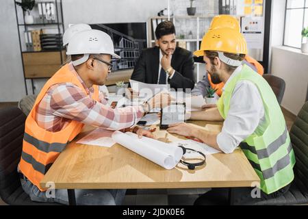 Multicultural workers in reflective vest and helmets using tablet for creating new plan of construction. Architects, designers and engineers leading conference at office. Brainstorming concept. Stock Photo