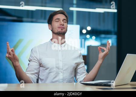 Young handsome man freelancer, businessman, manager in the office at the table resting, closed his eyes, meditates, shows his hands yoga poses. Stock Photo