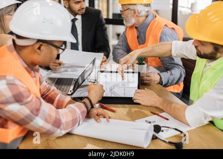 Close up of construction team analysing blueprints during business meeting at office. Multiracial partners using digital tablet for work. Concept of cooperation and brainstorming. Stock Photo