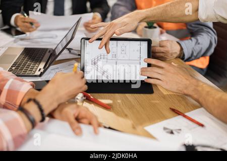 Close up of construction team analysing blueprints during business meeting at office. Multiracial partners using digital tablet for work. Concept of cooperation and brainstorming. Stock Photo