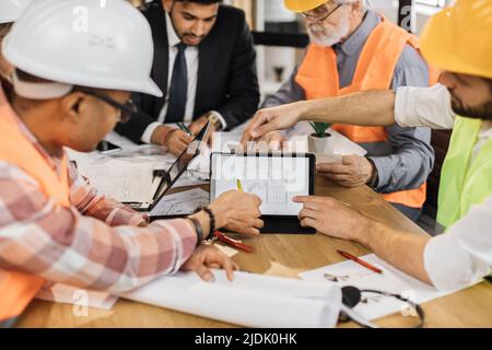 Close up of construction team analysing blueprints during business meeting at office. Multiracial partners using digital tablet for work. Concept of cooperation and brainstorming. Stock Photo
