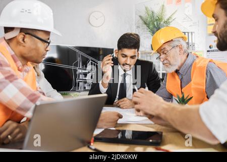 Architects and engineers in suits and helmets having working meeting at modern office. Colleagues of different ages and races. Corporate brainstorming. Stock Photo