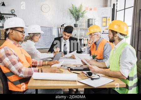 Multiracial men and women in formal outfit and helmets sitting at table with lots of blueprints during important strategic session. Business people working on common construction project. Stock Photo