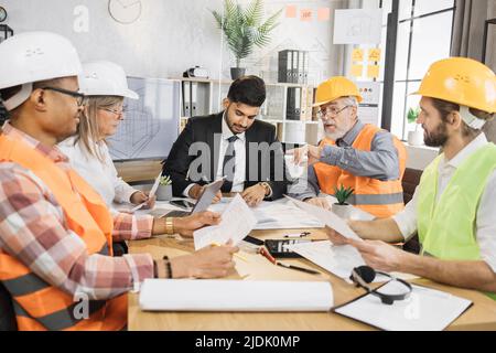 Multiracial men and women in formal outfit and helmets sitting at table with lots of blueprints during important strategic session. Business people working on common construction project. Stock Photo