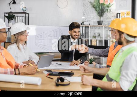 Multiethnic architects, designers and engineers sitting at table and greeting each other with hands shaking. Business partners having working meeting at office with big TV plasma screen. Stock Photo