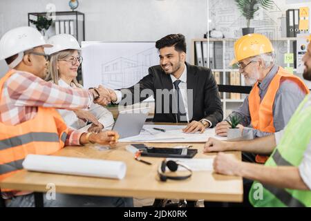 Multiethnic architects, designers and engineers sitting at table and greeting each other with hands shaking. Business partners having working meeting at office with big TV plasma screen. Stock Photo