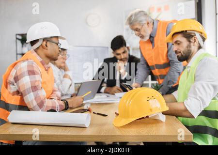 Focuse on protective hardhat. Competent engineers, designers and architects leading working conference at office center. Group of multicultural people wearing suit, uniform and protective helmet. Stock Photo