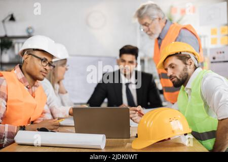 Group of coworkers in suits and helmets having business meeting at office. Responsible engineers focusing on common project of new building. Construction industry. Stock Photo