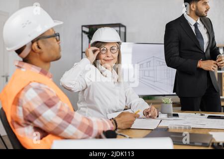 Beautiful senior woman in formal outfit sitting at table with males architects in uniform and helmet during office meeting. Group of people discussing construction project indoors. Stock Photo