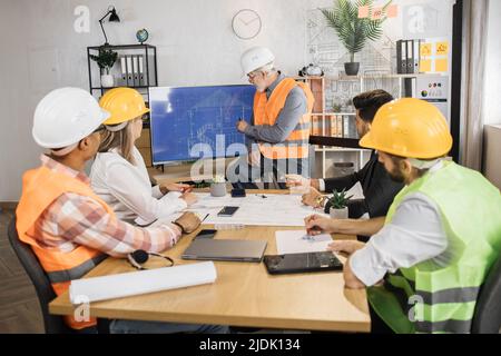 Senior male builder in hard hat showing drawings of new building on large digital screen. Group of five international partners having working meeting to discuss design and measurements of new building Stock Photo