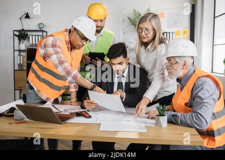 Group architects and engineers sitting at office and working on common project. Indian boss man in business suit sitting at the table and discusses construction strategy with colleagues and partners. Stock Photo