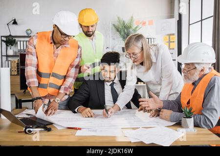 Indian man in suit presenting new construction project to colleagues in helmets and uniform at office. Three men and one woman standing near table and listening their competent partner. Stock Photo