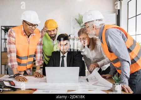 Four men and one woman sitting together at table and discussing blueprints on laptop during business conference. Multi ethnic architects, designers and engineers working at office. Stock Photo