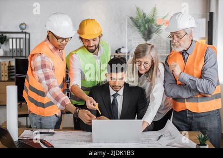Teams of architects, builders and designers working on project of new modern house. The head of company sits at table and shows on laptop sketches of building while contractors standing around. Stock Photo