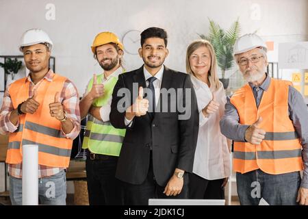 Front view of one senior woman and four men engineers, builders, architects looking at camera while standing on light modern office. People of different age, gender and race. Stock Photo