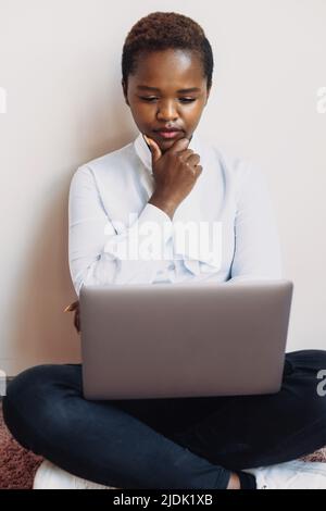 Pensive afro freelancer woman, looking at laptop screen while working remotely at home, sitting on floor. Black portrait. Internet technology Stock Photo
