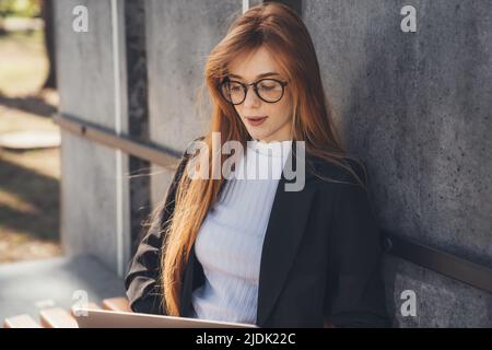 Beautiful young ginger redheaded woman in eyeglasses sitting on bench using laptop in the summer park. Internet technology. Online education. Stock Photo
