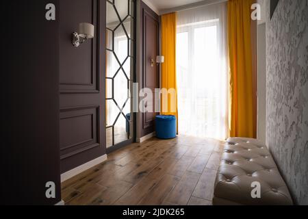 Modern entrance hall or hallway in residential building with large window and curtains, mirror, built-in wardrobe and sofa.