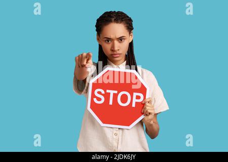 Portrait of serious woman with dreadlocks holding big red stop road sign and pointing finger on you, strictly looking at camera, wearing white shirt. Indoor studio shot isolated on blue background.