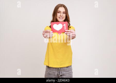 Portrait of beautiful teenager girl with brown hair showing social media heart Like icon, symbol of popularity in internet, recommending to push button. Indoor studio shot isolated on gray background. Stock Photo