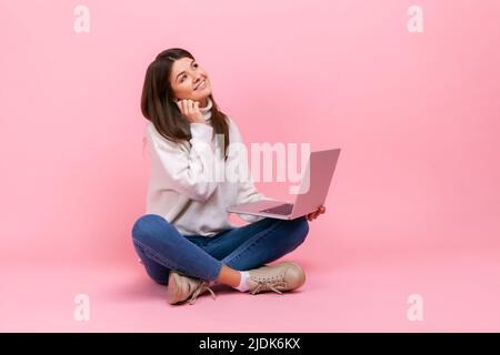 Portrait of female with dreamy facial expression, sitting with crossed legs on floor, holding laptop, wearing white casual style sweater. Indoor studio shot isolated on pink background. Stock Photo