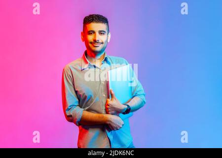 Portrait of smiling positive man in shirt holding folded laptop, looking at camera with happy facial expression, finish work. Indoor studio shot isolated on colorful neon light background. Stock Photo