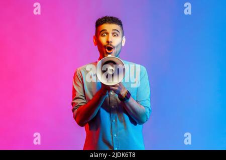 Portrait of astonished man in shirt holding megaphone in hands and looking at camera with shocked facial expression. Indoor studio shot isolated on colorful neon light background. Stock Photo