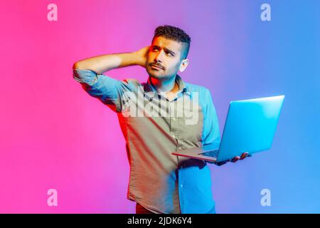 Portrait of handsome pensive man in shirt holding laptop and rubbing head looking away, thinking over new business project. Indoor studio shot isolated on colorful neon light background. Stock Photo