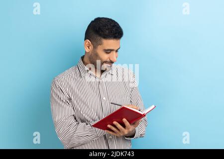 Smiling satisfied businessman with beard writing down to do list, making notes with pen in notebook, checking schedule, wearing striped shirt. Indoor studio shot isolated on blue background. Stock Photo