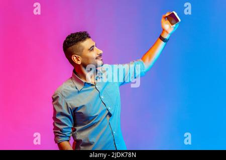 Portrait of positive optimistic man in shirt talking on video call, having online conversation on mobile phone, taking selfie. Indoor studio shot isolated on colorful neon light background. Stock Photo