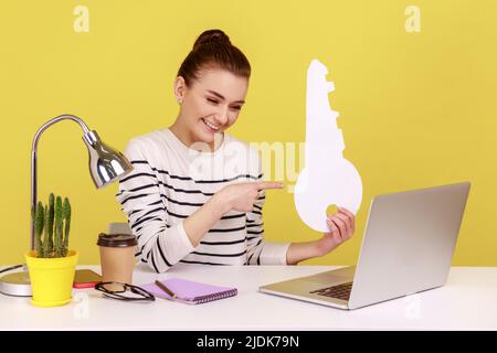 Woman office worker pointing at big paper key and looking at laptop screen with toothy smile while sitting on workplace, house purchase concept. Indoor studio studio shot isolated on yellow background Stock Photo