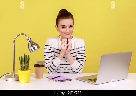 Cunning young adult woman with tricky face scheming and conspiring, pondering devious sly business plan while sitting on workplace. Indoor studio studio shot isolated on yellow background. Stock Photo