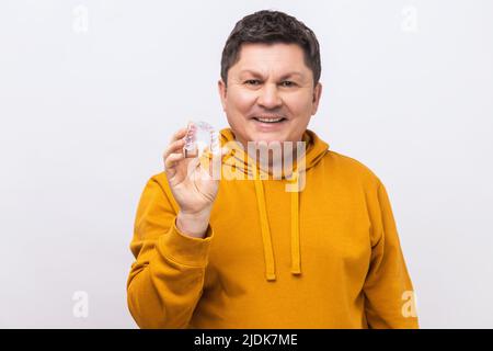 Portrait of smiling middle aged man with dark hair holding transparent plastic braces, mouth guard, wearing urban style hoodie. Indoor studio shot isolated on white background. Stock Photo