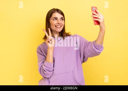 Portrait of optimistic happy woman waving hand to smartphone and taking selfie, showing v sign while making video call, wearing purple hoodie. Indoor studio shot isolated on yellow background. Stock Photo