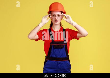 Portrait of confident builder woman standing putting on protective glasses, looking at camera, wearing overalls and protective helmet. Indoor studio shot isolated on yellow background. Stock Photo