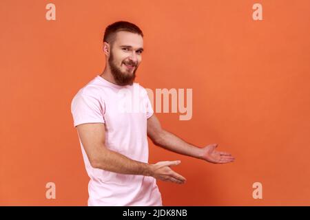 Portrait of kind-hearted generous bearded man showing welcome gesture and empty space on wall for your best advertising, wearing pink T-shirt. Indoor studio shot isolated on orange background. Stock Photo