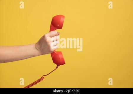 Profile side view closeup of woman hand holding and showing red call telephone handset receiver. Indoor studio shot isolated on yellow background. Stock Photo