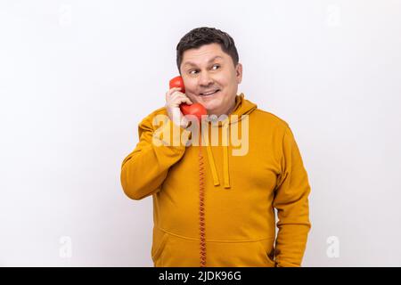 Happy middle aged man talking red landline telephone, holding in hand handset, looking away with toothy smile, wearing urban style hoodie. Indoor studio shot isolated on white background. Stock Photo