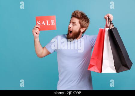 Portrait of excited bearded man holding paper bags and looking at sale caer, shopping and discounts, yelling happily, expressing amazement. Indoor studio shot isolated on blue background. Stock Photo